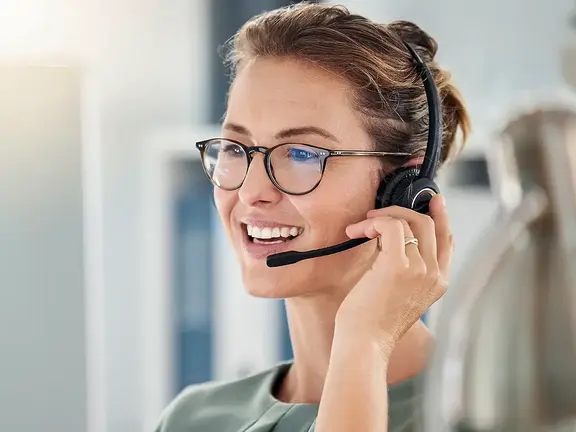 Lächelnde Frau, die eine Brille und ein Headset trägt, spricht und arbeitet in einer hellen Büroumgebung.