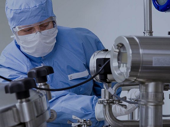 Eine Person in Schutzkleidung bedient Geräte aus rostfreiem Stahl in einem sterilen Labor oder einer pharmazeutischen Einrichtung.