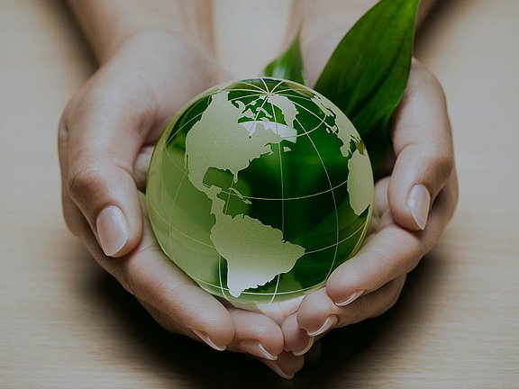 Zwei Hände halten sanft einen grünen Globus mit sichtbaren Kontinenten, daneben ein grünes Blatt, das Umweltschutz symbolisiert.